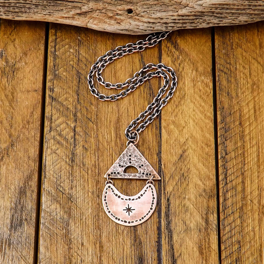 Full view of a copper necklace with a cut out sun design within a textured triangle that is attached with jump rings above a crescent moon with a cut out star burst in the center.