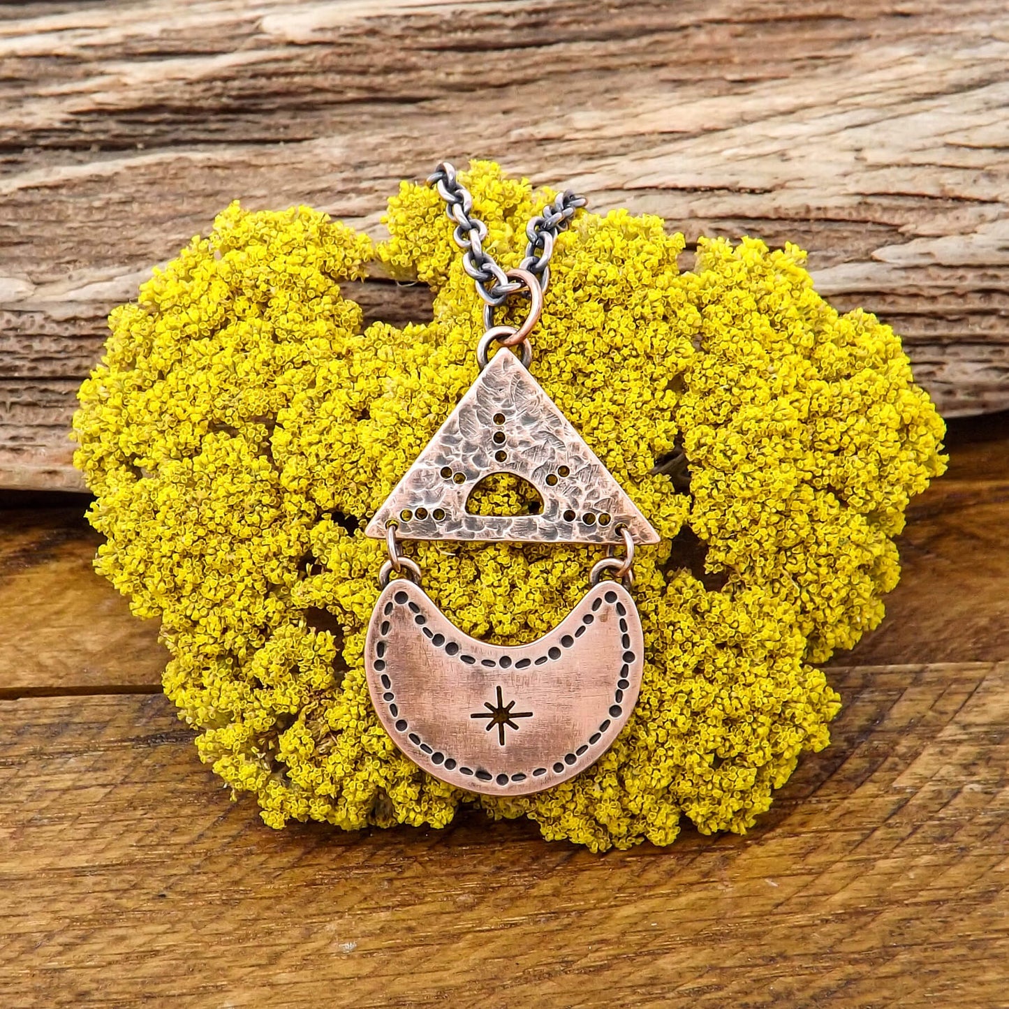 Front view of a copper necklace with a cut out sun design within a textured triangle that is attached with jumps rings above a crescent moon with a cut out star burst design in the center.