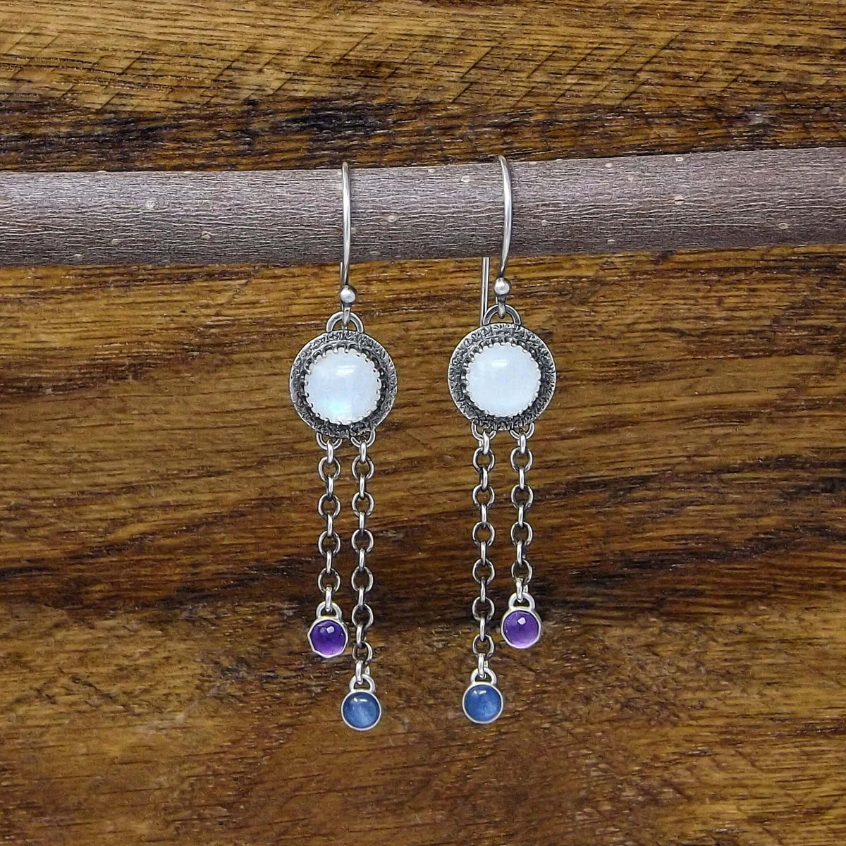 Front view of a pair of sterling silver dangle earrings featuring rainbow moonstone, kyanite, and rose cut faceted amethyst gemstones. Heart designs adorn the back of the earrings.