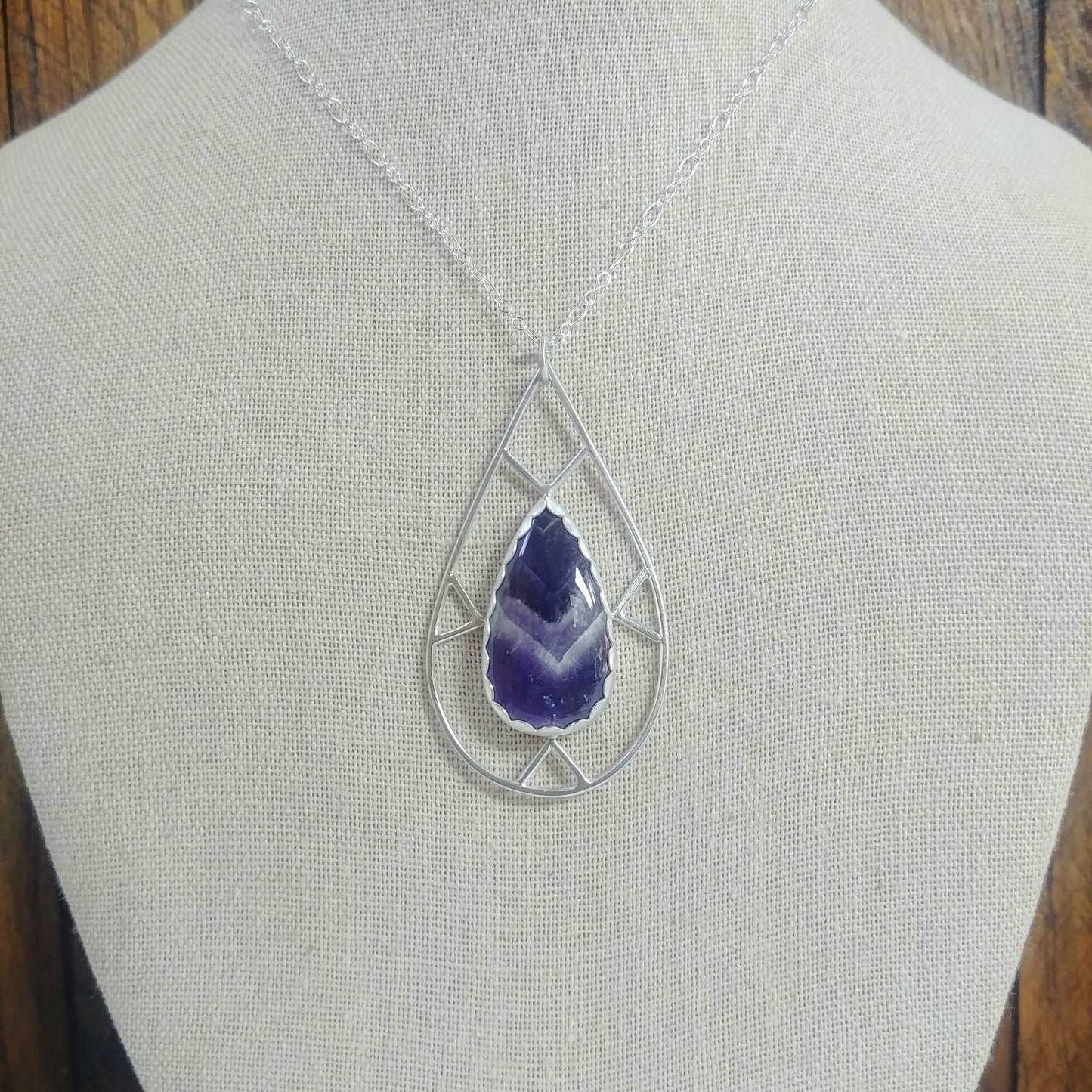 Display bust view of an argentium silver necklace with a geometric design and featuring a teardrop shaped chevron amethyst gemstone.