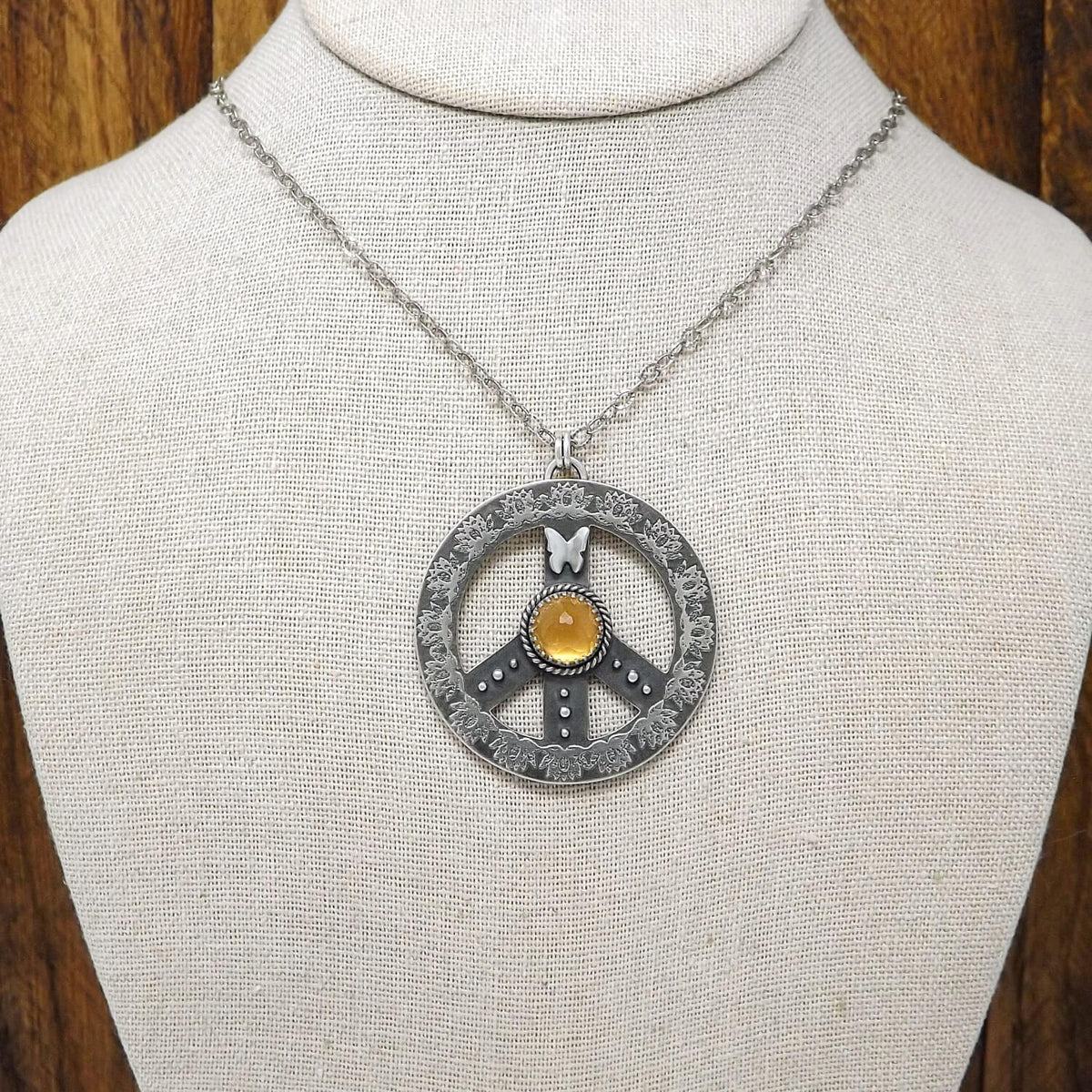 Display bust view of a sterling silver peace sign necklace with etched lotus flowers, a butterfly, argentium silver granules and a rose cut faceted hessonite garnet.