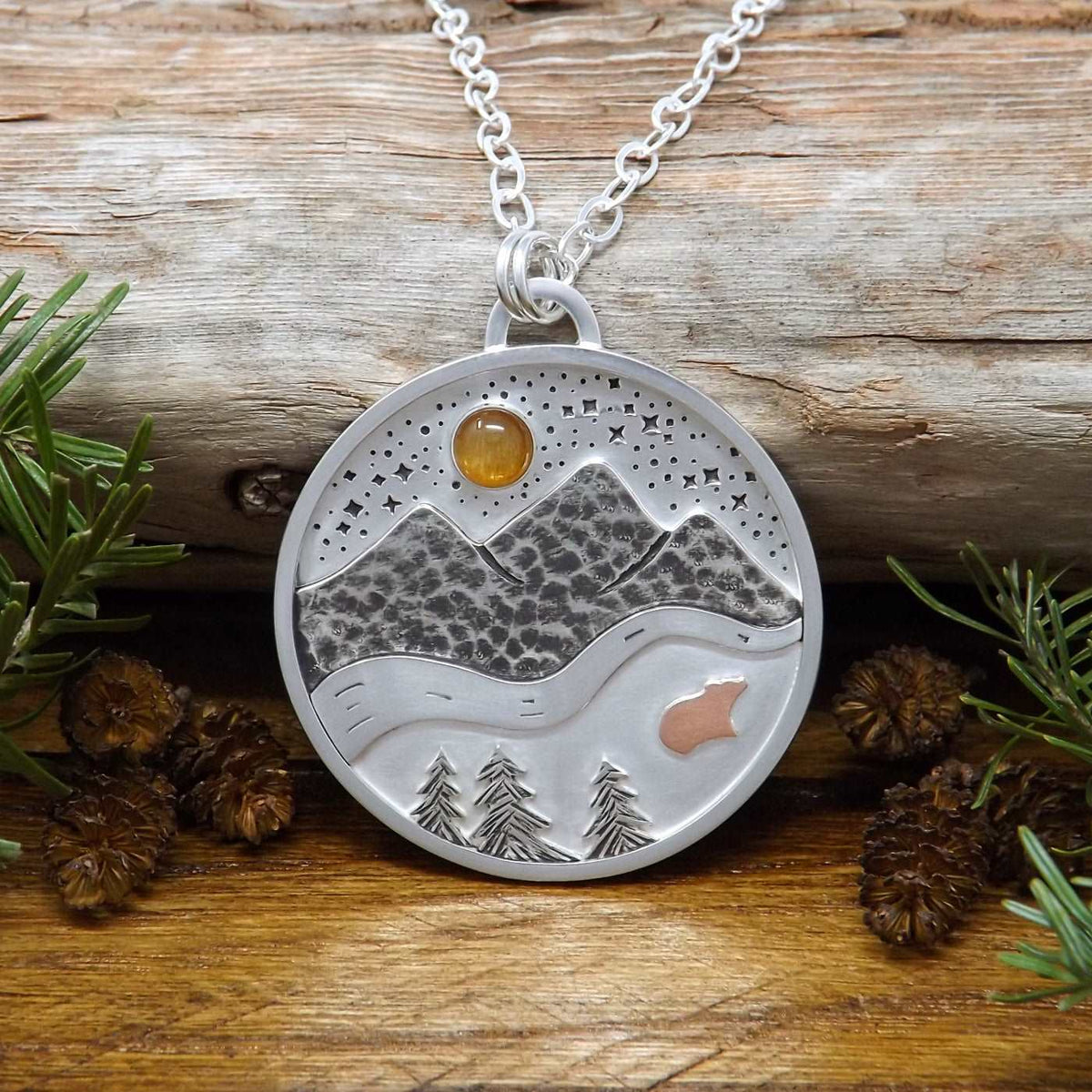 Front view of a sterling silver necklace with nature scenery including a gold sunstone gemstone in a starry night sky, mountains, a river, trees, and a tiny copper bear.