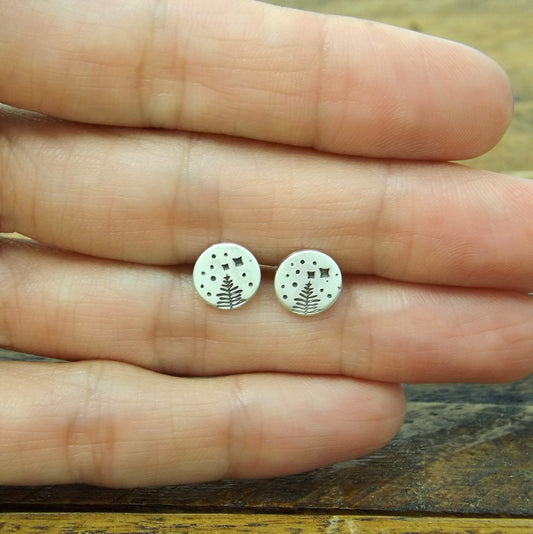 Size reference view of a pair of sterling silver stud earrings with a hand-stamped starry night pine tree design.