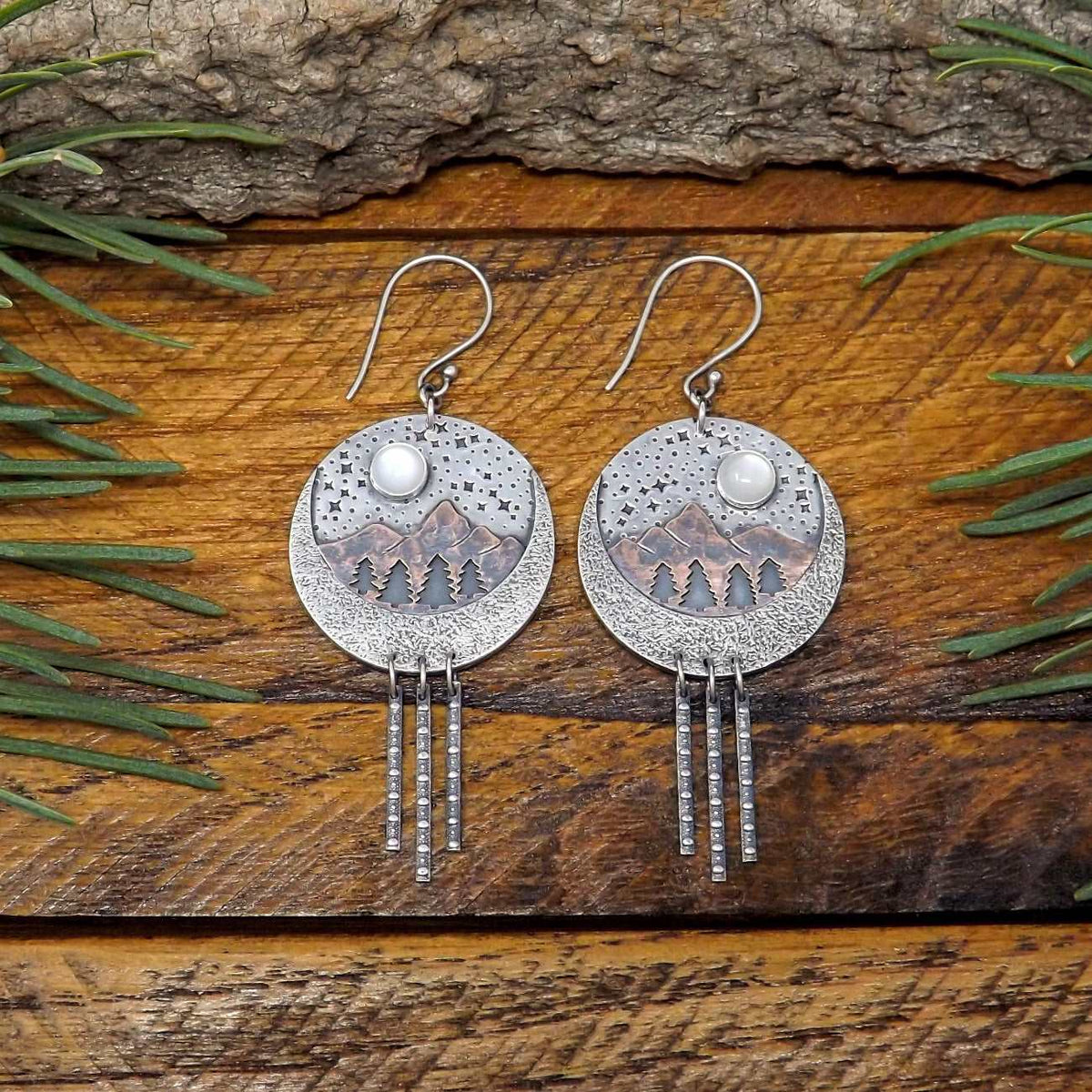Front view of a pair of sterling silver and copper dangle earrings with a starry night forest and mountain scenery. Moonstone gemstones adorn the starry night sky.