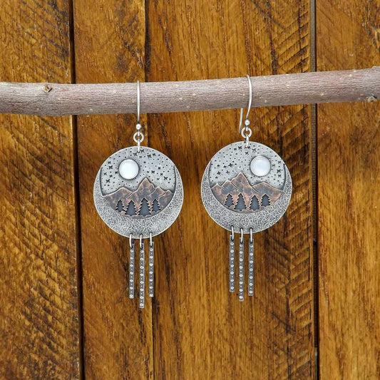 Hanging view of a pair of sterling silver and copper dangle earrings with a starry night forest and mountain scenery. Moonstone gemstones adorn the starry night sky.