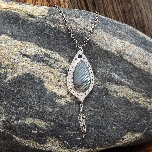 Left angle view of a sterling silver necklace with a teardrop shaped labradorite gemstone that is surrounded by hand-stamped circle details. Dangling below is a feather charm.