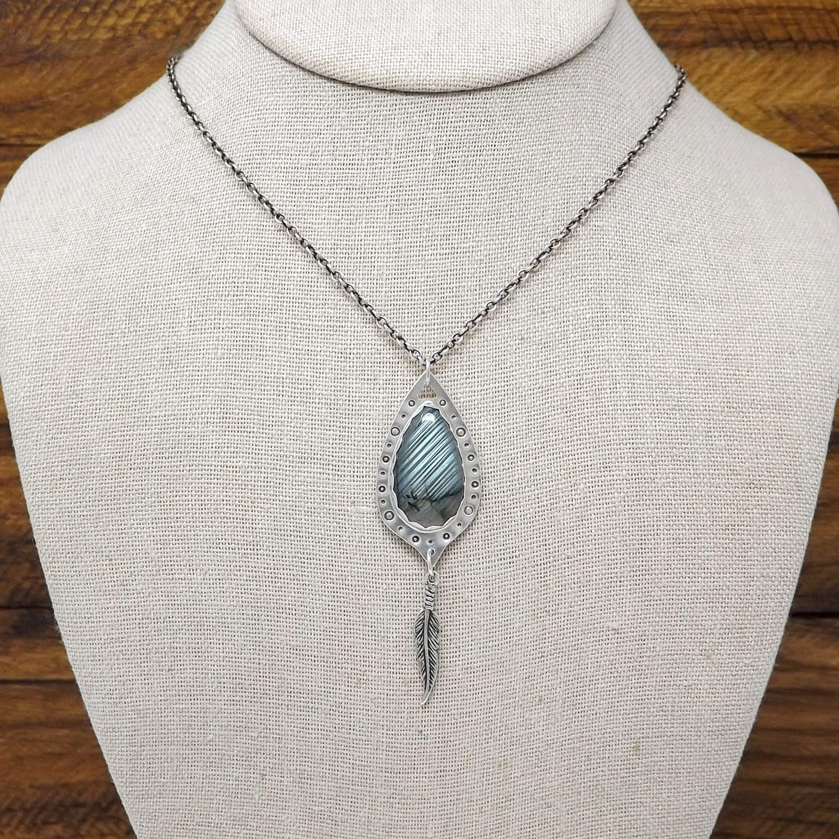 Display bust view of a sterling silver necklace with a teardrop shaped labradorite gemstone that is surrounded by hand-stamped circle details. Dangling below is a feather charm.