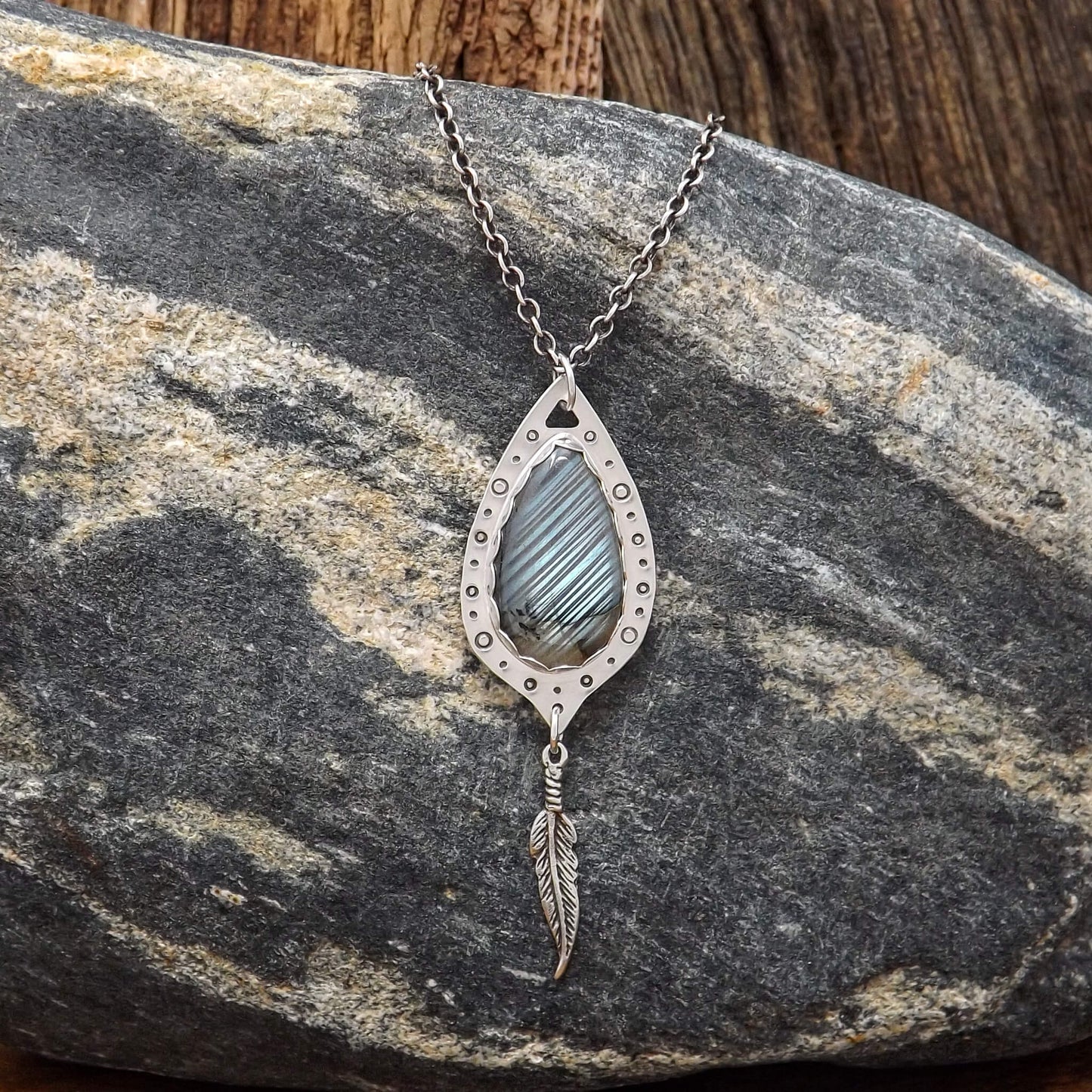 Front view of a sterling silver necklace with a teardrop shaped labradorite gemstone that is surrounded by hand-stamped circle details. Dangling below is a feather charm.