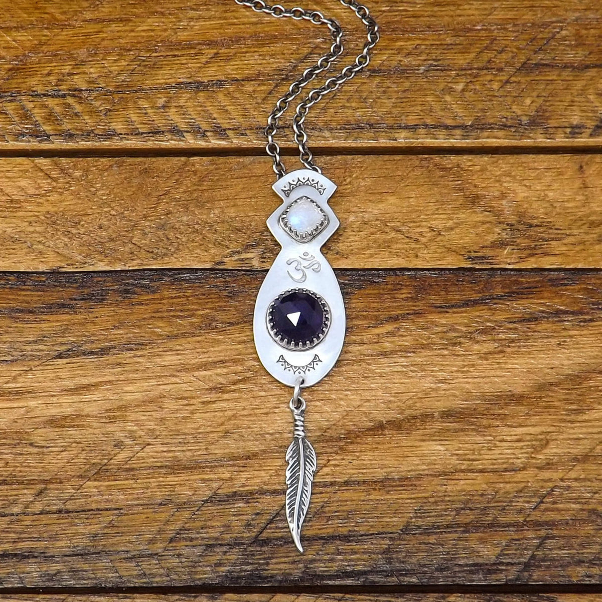Front view of a sterling silver necklace with a hand-stamped Om detail in the center. Also featuring a faceted square rainbow moonstone and rose cut faceted amethyst gemstone. At the bottom of the pendant is a feather charm.