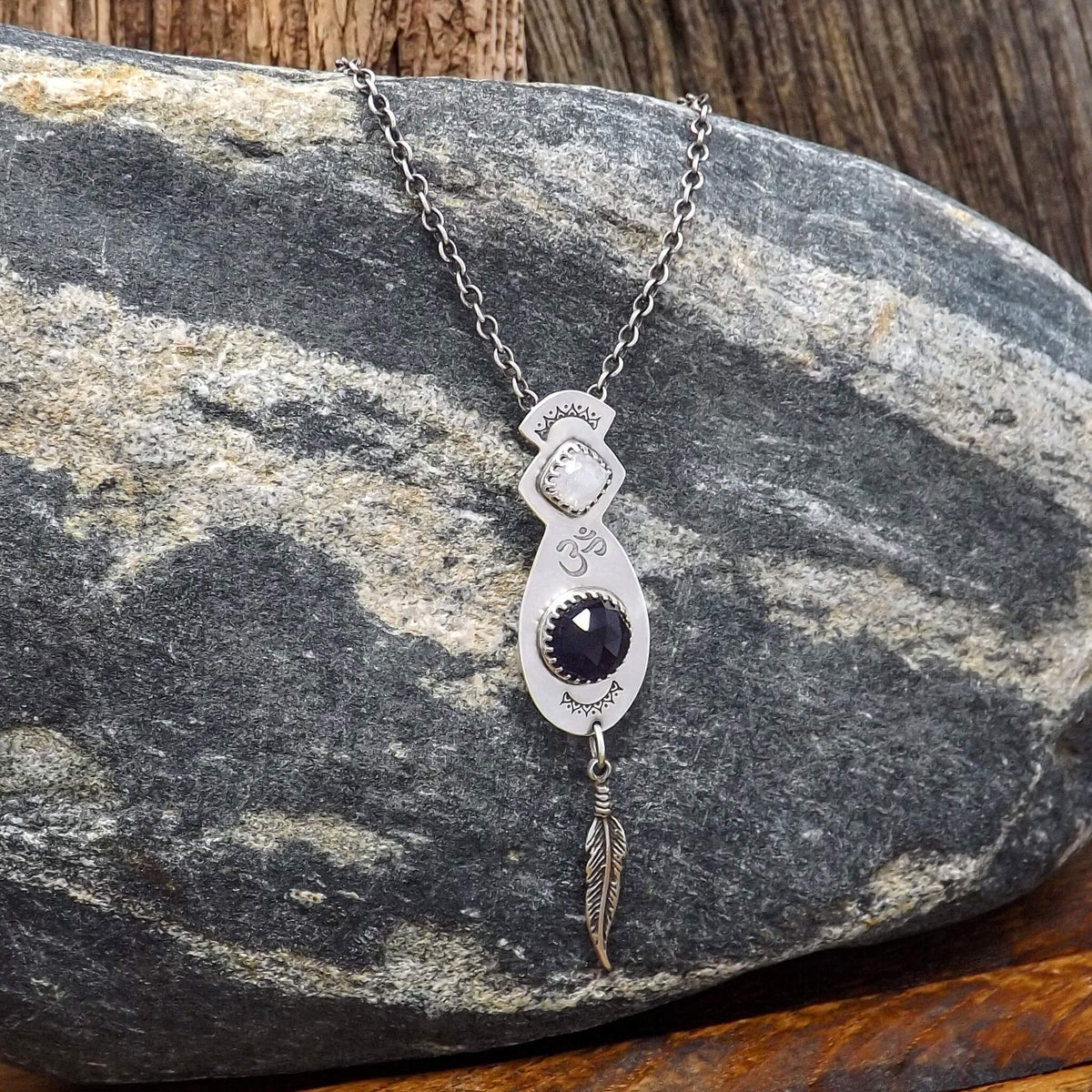 Left angle view of a sterling silver necklace with a hand-stamped Om detail in the center. Featuring a square faceted rainbow moonstone and rose cut faceted amethyst gemstone. At the bottom of the pendant is a feather charm.