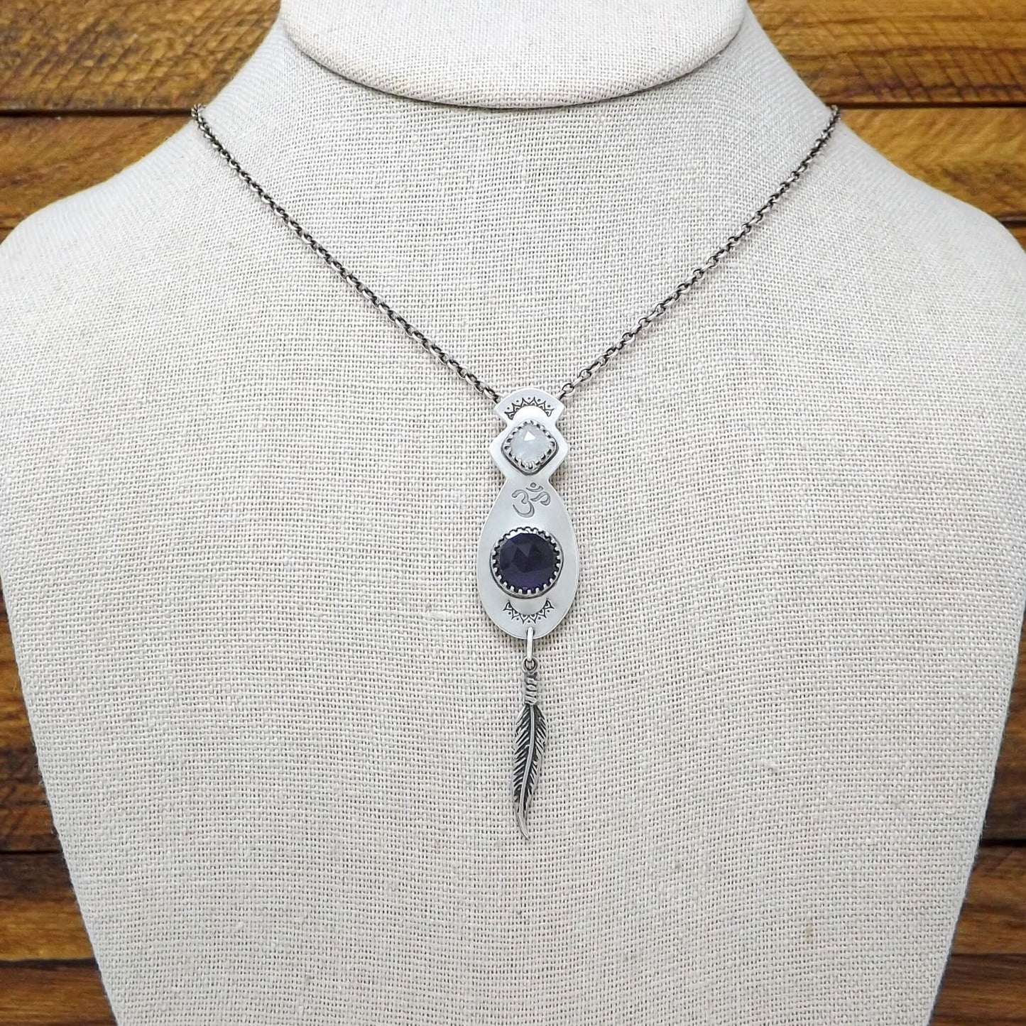Display bust view of a sterling silver necklace with a hand-stamped Om detail in the center. Featuring a square faceted rainbow moonstone and rose cut faceted amethyst gemstone. At the bottom of the pendant is a feather charm.