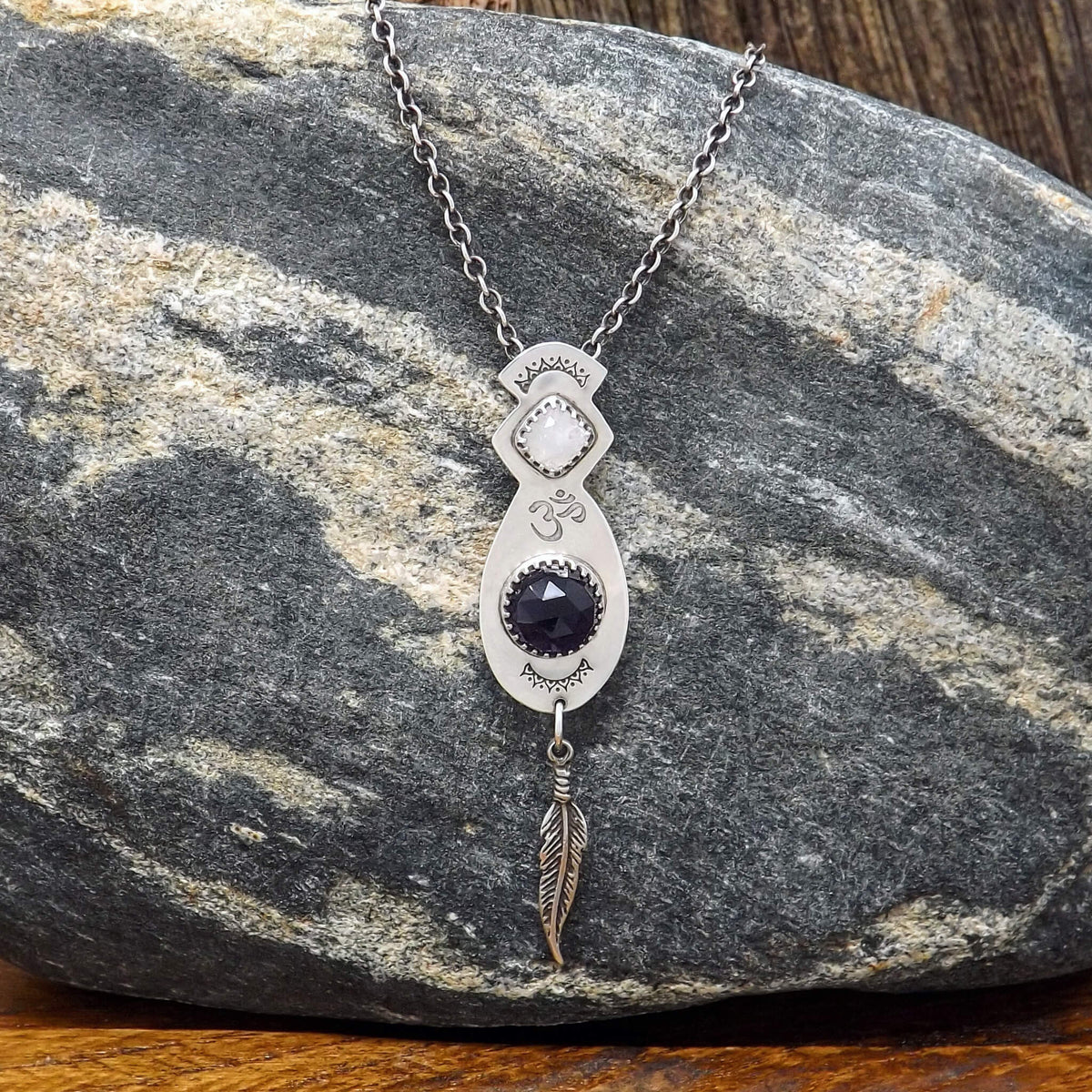 Close up view of a sterling silver necklace with a hand-stamped Om detail in the center. Featuring a square faceted moonstone and rose cut faceted amethyst gemstone. At the bottom of the pendant is a feather charm.