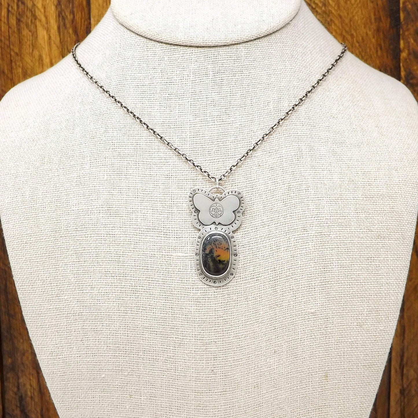Bust view of a sterling silver dendritic agate gemstone necklace adorned with a butterfly stamped with the Jenn's Zen Creations logo. A hand-stamped design borders the pendant.