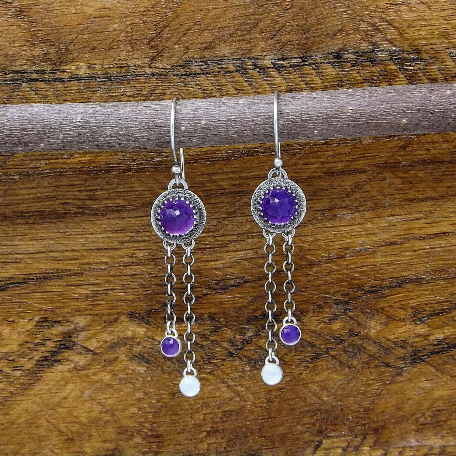 Front view of a pair of sterling silver dangle earrings featuring rose cut faceted amethyst, and rainbow moonstone gemstones. Heart designs adorn the back of the earrings.