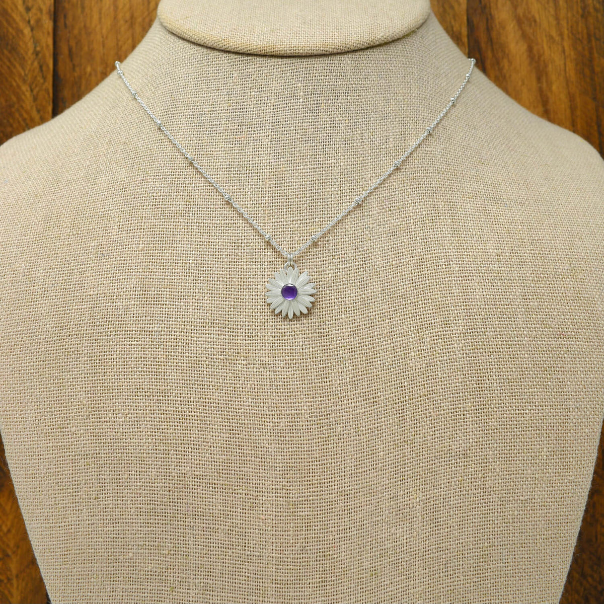 A sterling silver daisy necklace with a faceted amethyst at the flower center, displayed on a necklace bust.