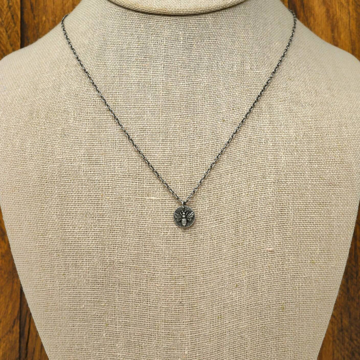 Hand-fabricated sterling silver heart necklace with a 3-dimensional bee on a hand-stamped textured disc, displayed on a necklace bust.